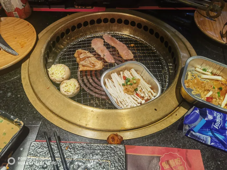 照片：烤肉店的圆桌，篦子上正烤着肉片，旁边有锡纸金针菇与配菜。