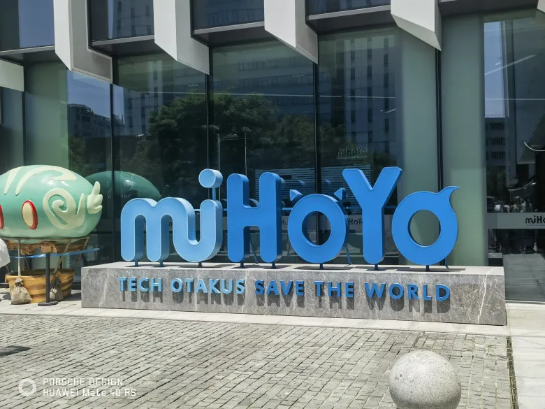 照片：米哈游公司门口的蓝色 miHoYo 立体招牌与「TECH OTAKUS SAVE THE WORLD」基座。
