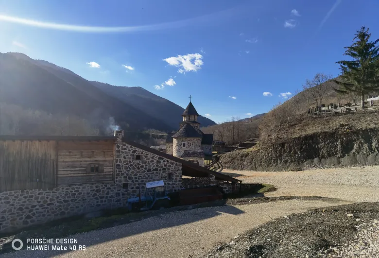 照片：山路旁的石砌小教堂，尖顶十字架与木屋相邻，远处是被阳光照亮的大山。