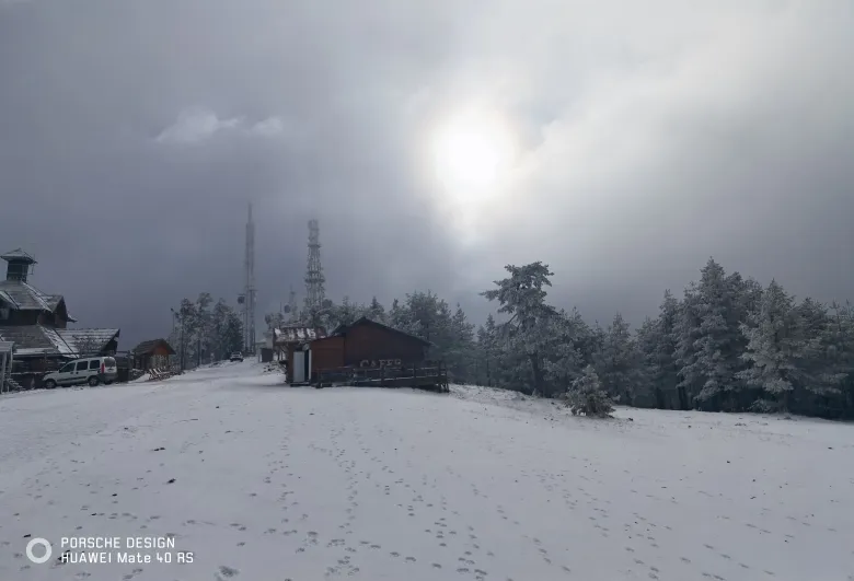 照片：积雪的山顶站，覆霜林木间有通信塔与小木屋，云缝中透出亮光。